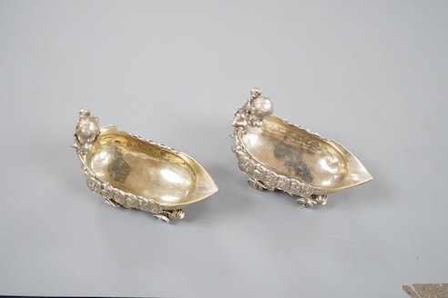 A pair of late 19th/early 20th century Hanau white metal salts, modelled as boat's with wheels and putti surmounts, maker Stork & Sinsheimer, length 81mm, 195 grams.